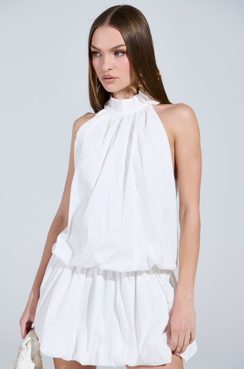A long-haired person in a chic, sleeveless, high-neck "Dressed for Any Occasion" white poplin mini dress with ruched waist and skirt holds a white textured bag against a plain gray background.
