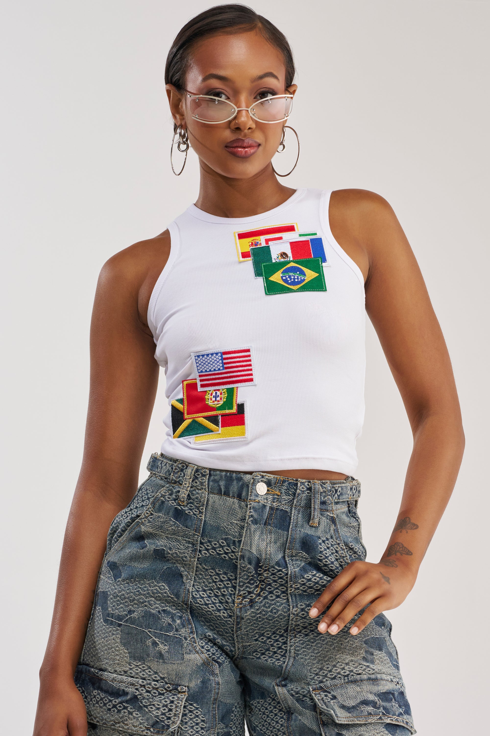 A woman wearing glasses models the AROUND THE WORLD TANK TOP—a cropped white streetwear tank embroidered with international flag patches (USA, Brazil, Italy, Portugal, and Spain)—paired with blue patterned jeans and posing confidently with one hand on her hip.