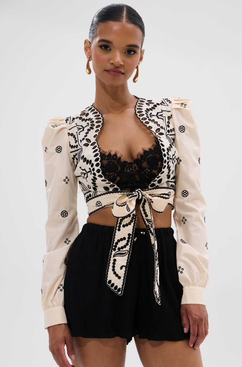 A woman wears the BELLINI FLORAL TIE FRONT TOP, a cream, long-sleeve, embroidered cropped jacket tied at the waist over a black lace bralette and high-waisted black shorts. She accessorizes with gold earrings and has slicked-back hair.