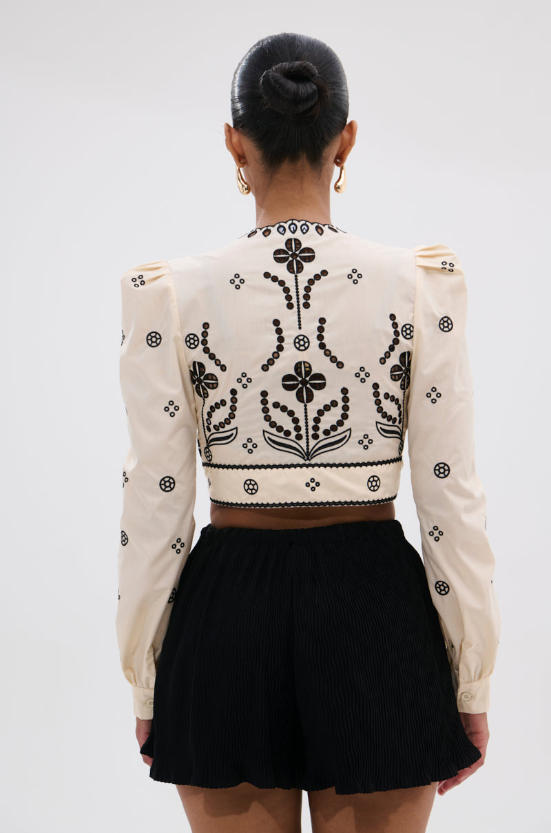 A woman with her hair in a bun is seen from the back, wearing the BELLINI FLORAL TIE FRONT TOP—a black floral blouse with cream long sleeves—paired with high-waisted black pleated shorts against a plain light gray background.