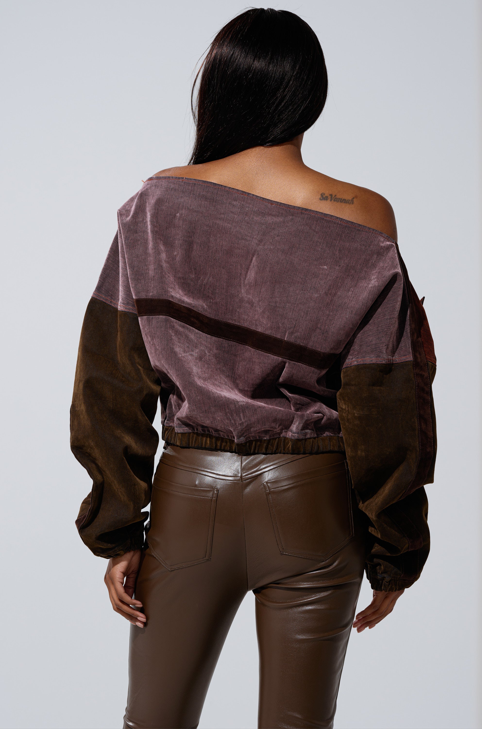 A woman with long dark hair, seen from behind, wears the PATCH ME UP MINERAL WASHED SWEATSHIRT IN PURPLE and brown leather pants. A small tattoo is visible on her upper right shoulder against a plain light background. 3