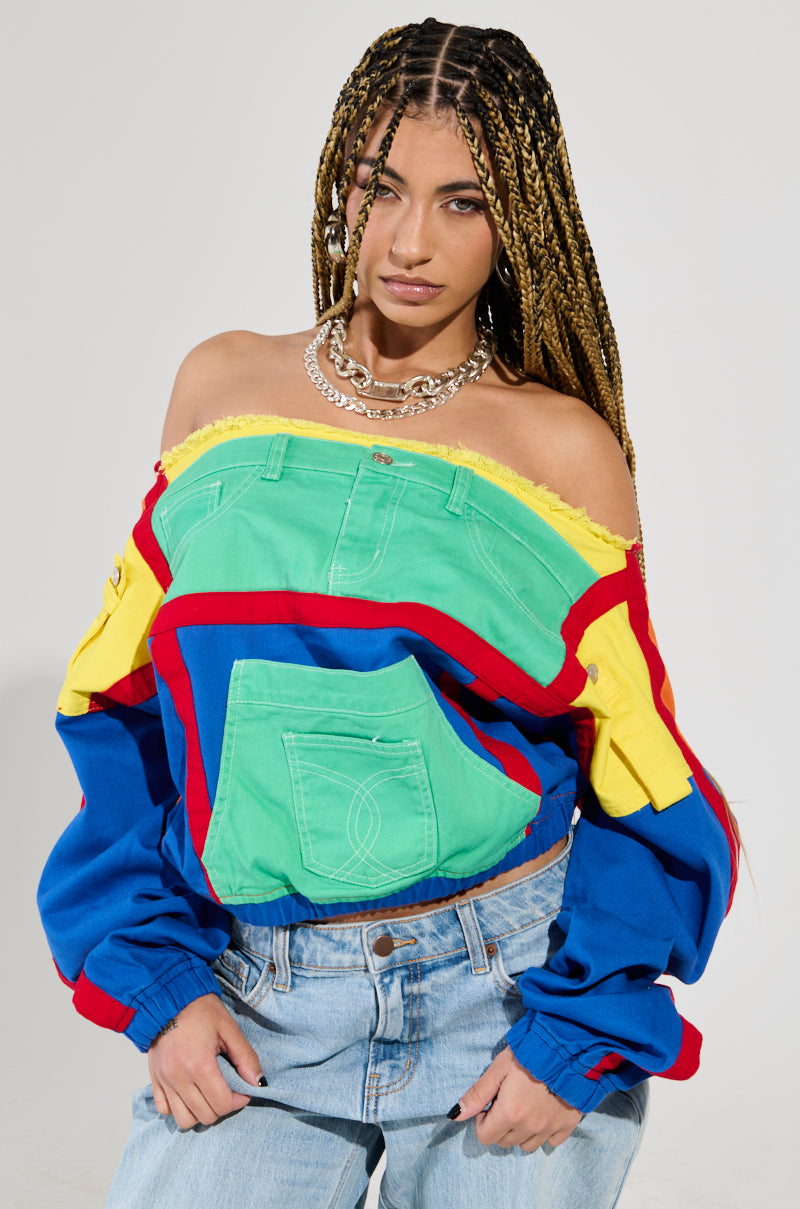 A person with long braided hair wears the PATCH ME UP SWEATSHIRT, showcasing an off-the-shoulder style with vibrant color block patches of red, green, blue, and yellow. They pair it with light denim jeans and accessorize with hoop earrings and layered necklaces against a plain gray background.