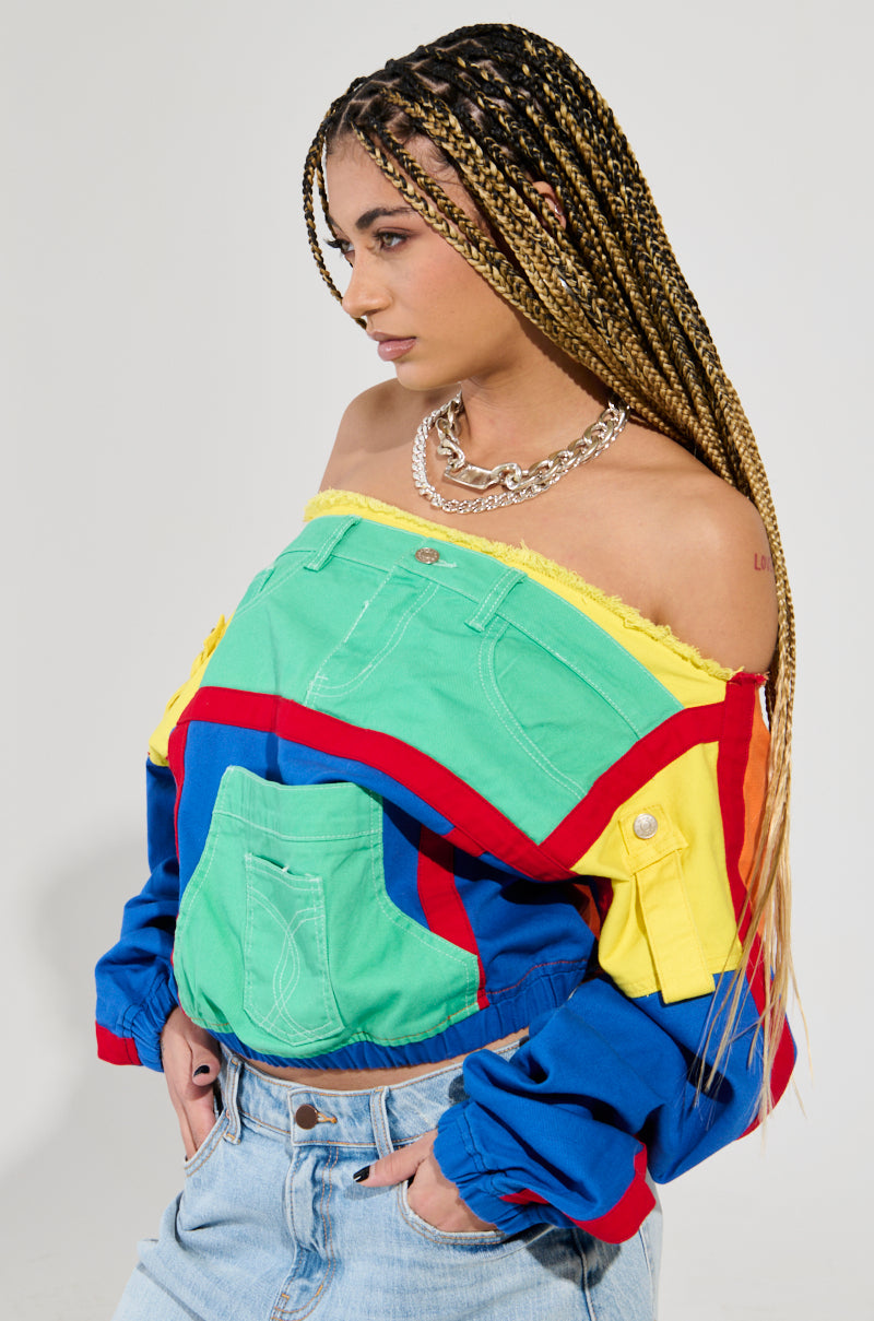 The individual with long braided hair is wearing the PATCH ME UP SWEATSHIRT, a colorful, off-the-shoulder top featuring vibrant green, yellow, red, and blue patchwork. They complete their look with light blue jeans and a silver necklace for an effortlessly chic style.