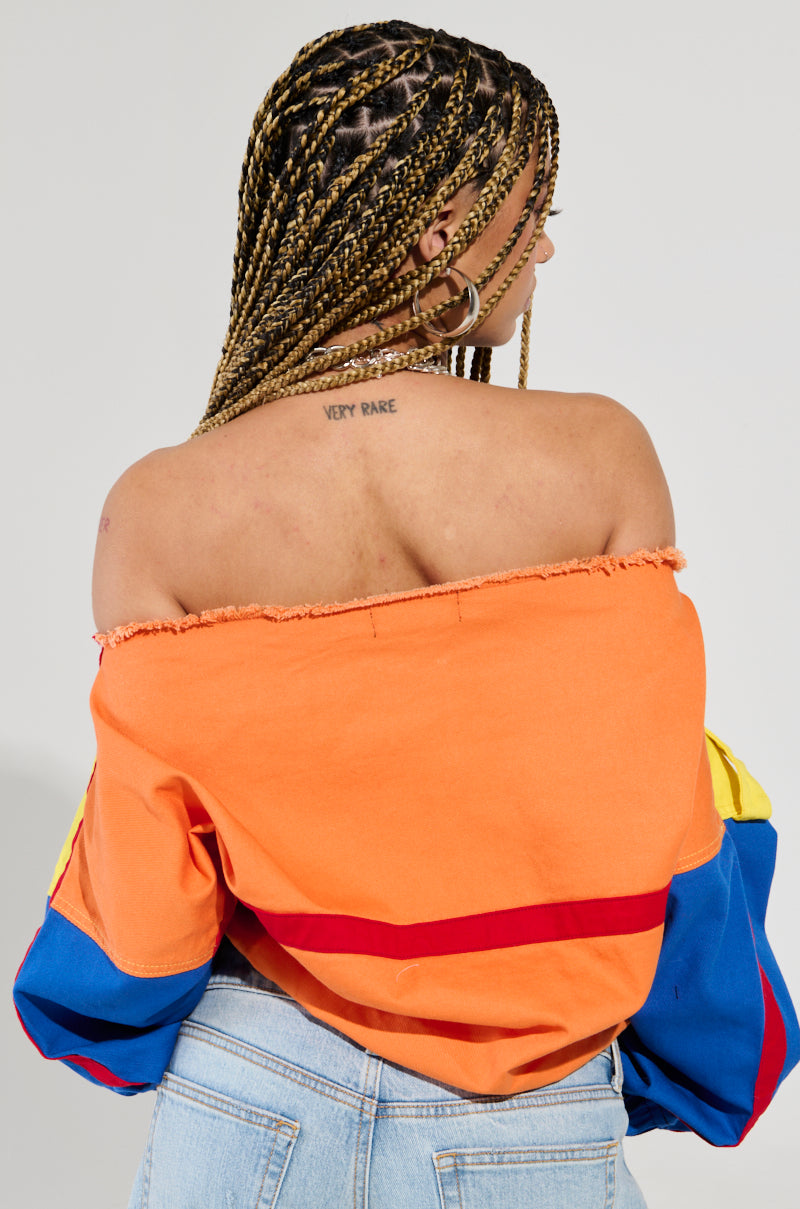 A person with braided hair is turned away, showcasing a PATCH ME UP SWEATSHIRT featuring vibrant orange, red, and blue colors. "Very Rare" is tattooed on their upper back. Hoop earrings dangle as they wear light blue jeans with an oversized kangaroo pocket for added flair.
