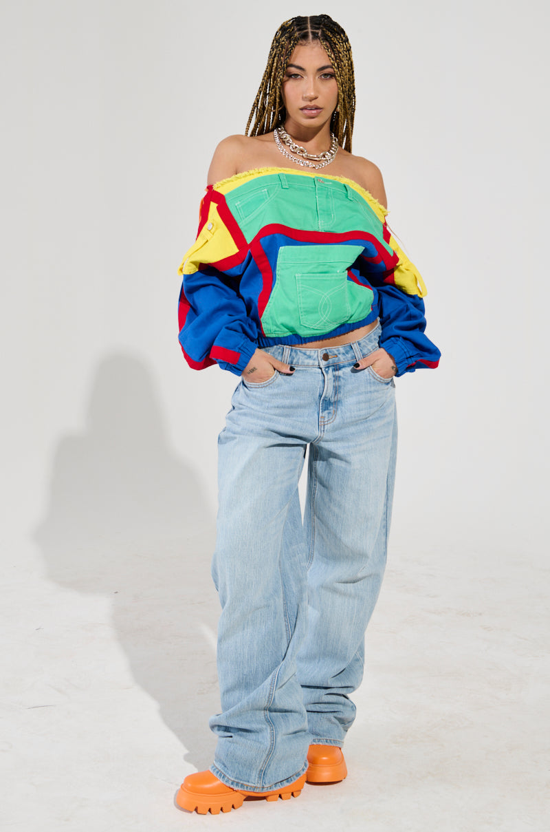 A person with braided hair stands confidently in the vibrant PATCH ME UP SWEATSHIRT, designed with an off-the-shoulder, color block pattern. They pair it stylishly with baggy light blue jeans, orange shoes, and a necklace, posing against a simple, light-colored background.