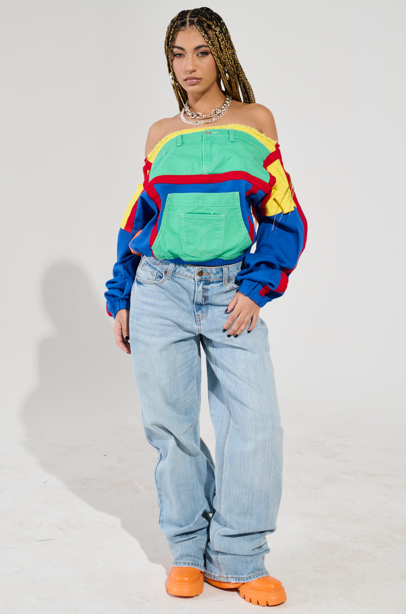 A person with braided hair wears the PATCH ME UP SWEATSHIRT, a color block off-the-shoulder piece featuring green, yellow, red, and blue hues. They pair it with light blue jeans and orange shoes while standing confidently against a plain background.