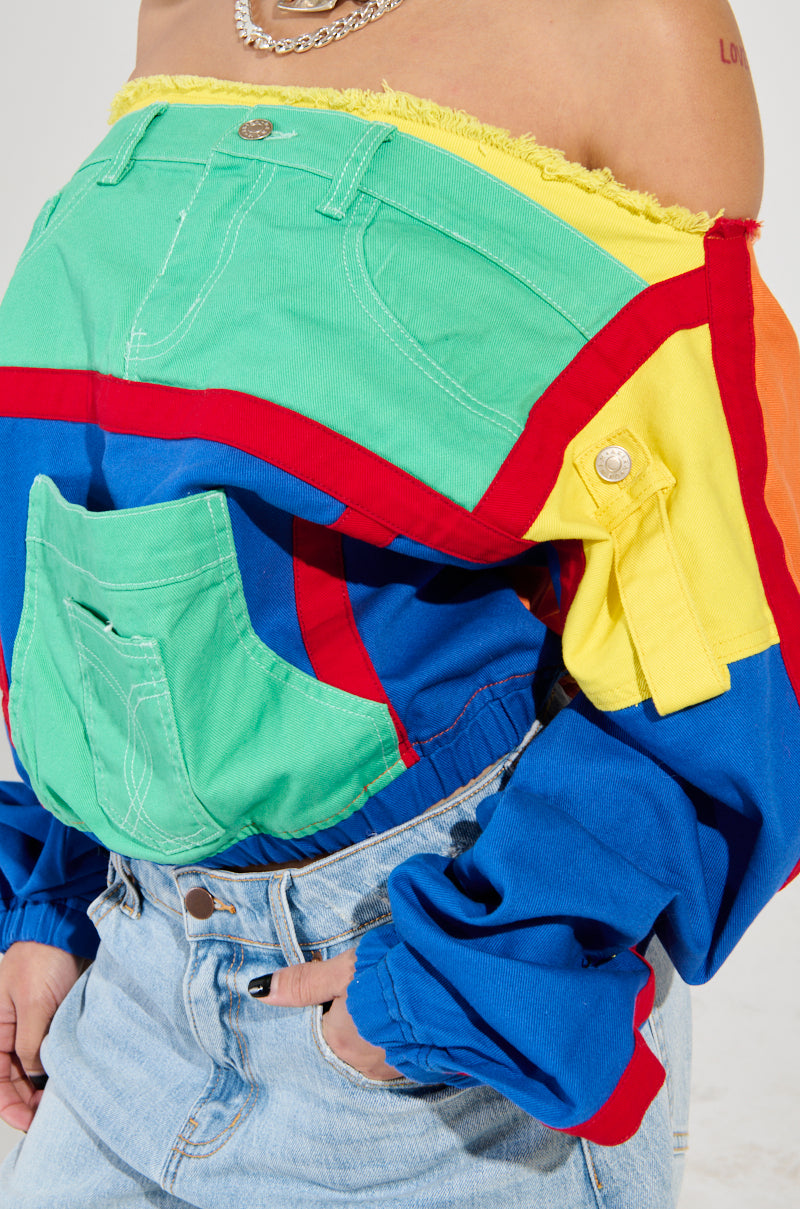 A person sporting the PATCH ME UP SWEATSHIRT, a vibrant off-the-shoulder top with a colorful patchwork design of jeans pockets in green, red, and yellow, pairs it flawlessly with light blue denim jeans. An oversized kangaroo pocket and chain necklace complete the outfit while highlighting a tattoo on their shoulder.
