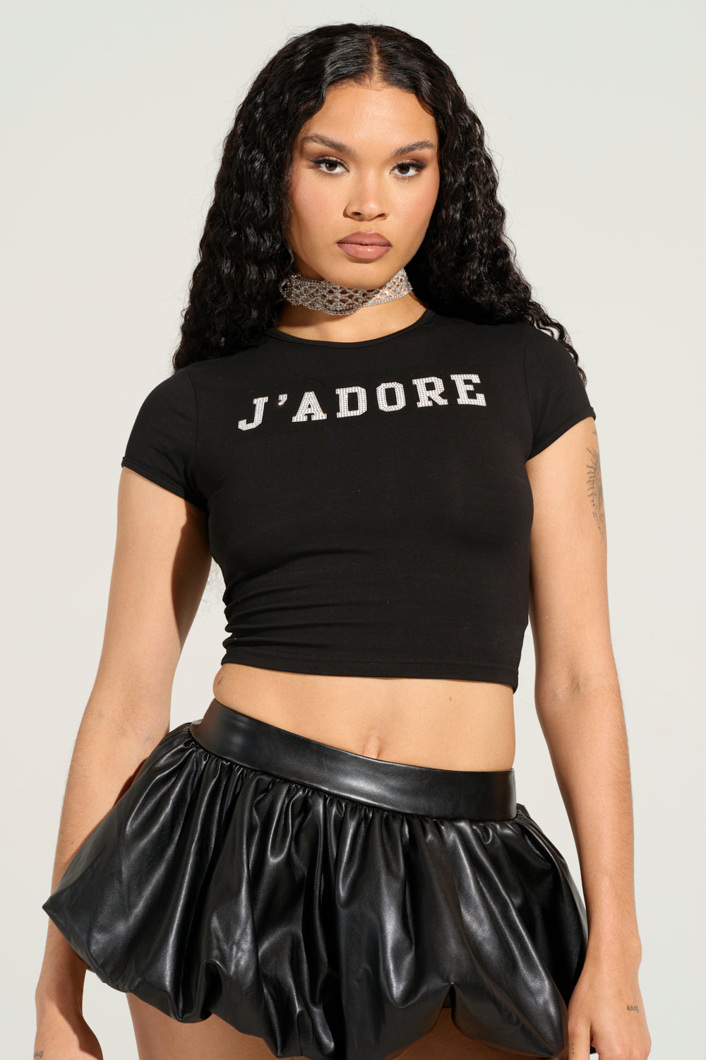 A person with long curly hair wears a black OUI MERCI BABY TEE from AKIRA, adorned with rhinestone details, paired with a shiny pleated black skirt. They accessorize with a silver choker and maintain a neutral expression while posing against a plain background.
