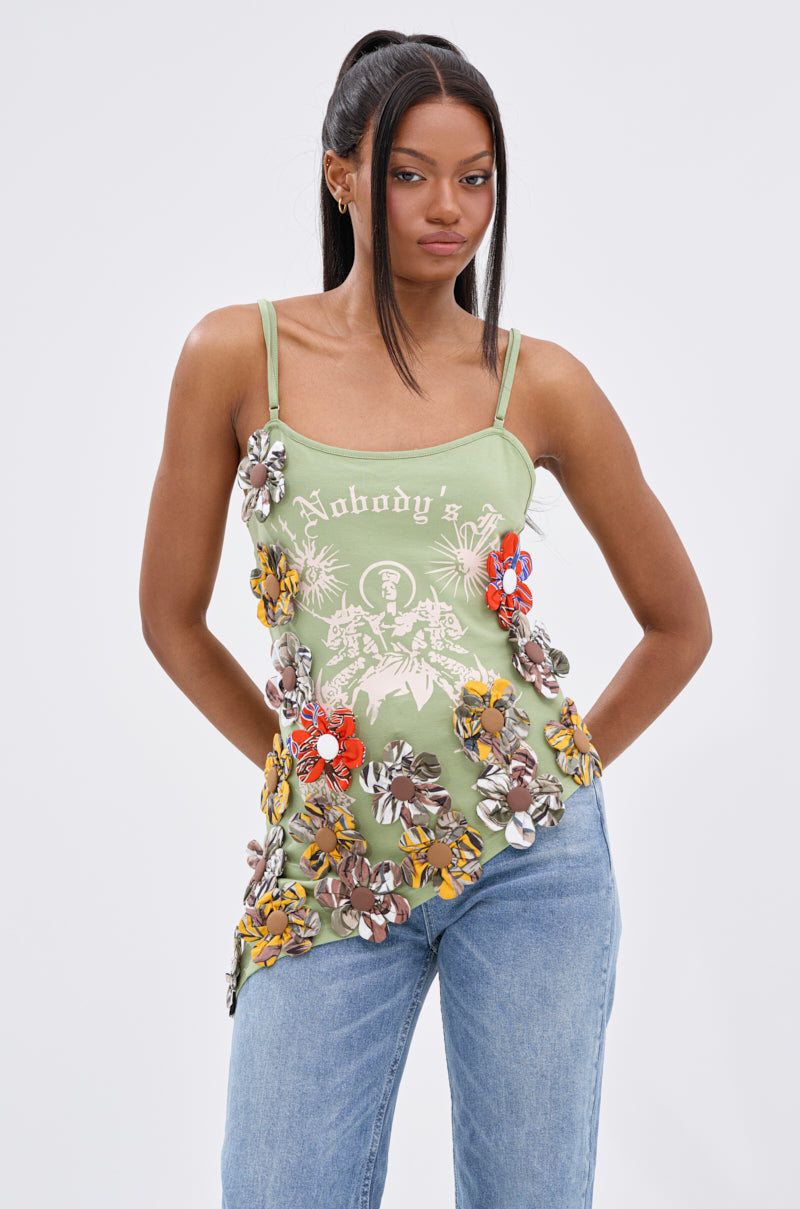 A woman wears the FOR THE FREE FLORAL EMBELLISHED TOP, a light green boho style with floral embellishments and "Nobody's" print, paired with blue jeans. Her long dark hair is half-up, half-down as she stands against a plain background.