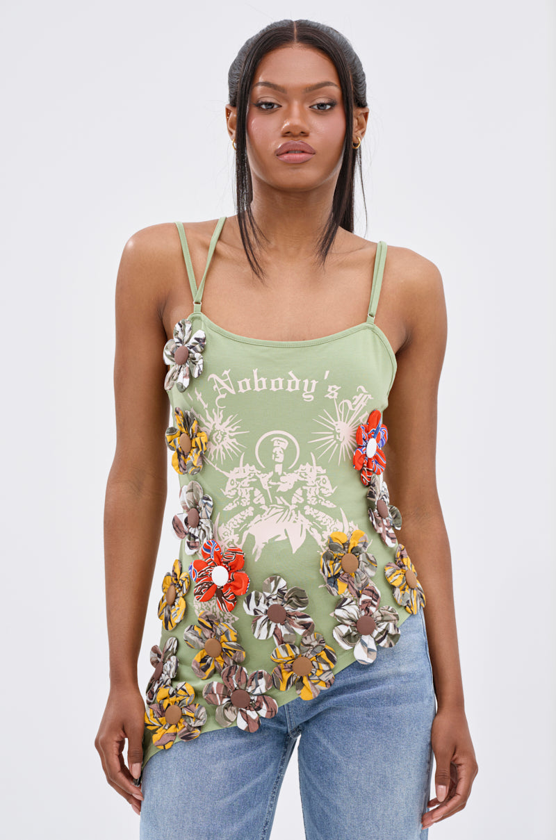 A woman stands against a plain background wearing the FOR THE FREE FLORAL EMBELLISHED TOP—a light green, stretchy cotton top with “Nobody’s” text, white graphic design, colorful 3D floral appliques—paired with light blue jeans.