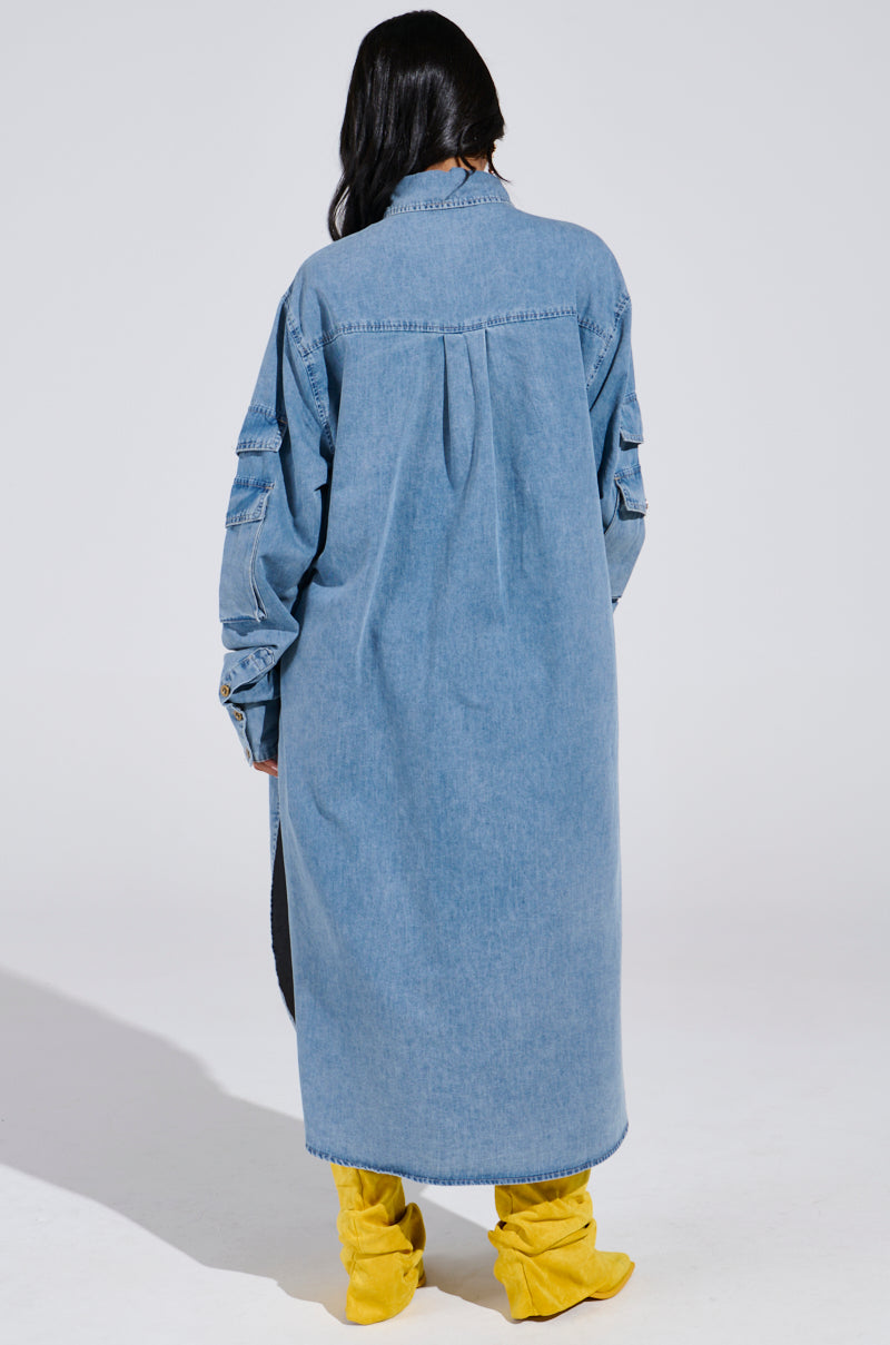 A person with long black hair, facing away, is wearing the COMFORT QUEEN CHAMBRAY LONG DUSTER and bright yellow slouchy boots. The simple light gray background accentuates the flowing maxi-length silhouette of their outfit.