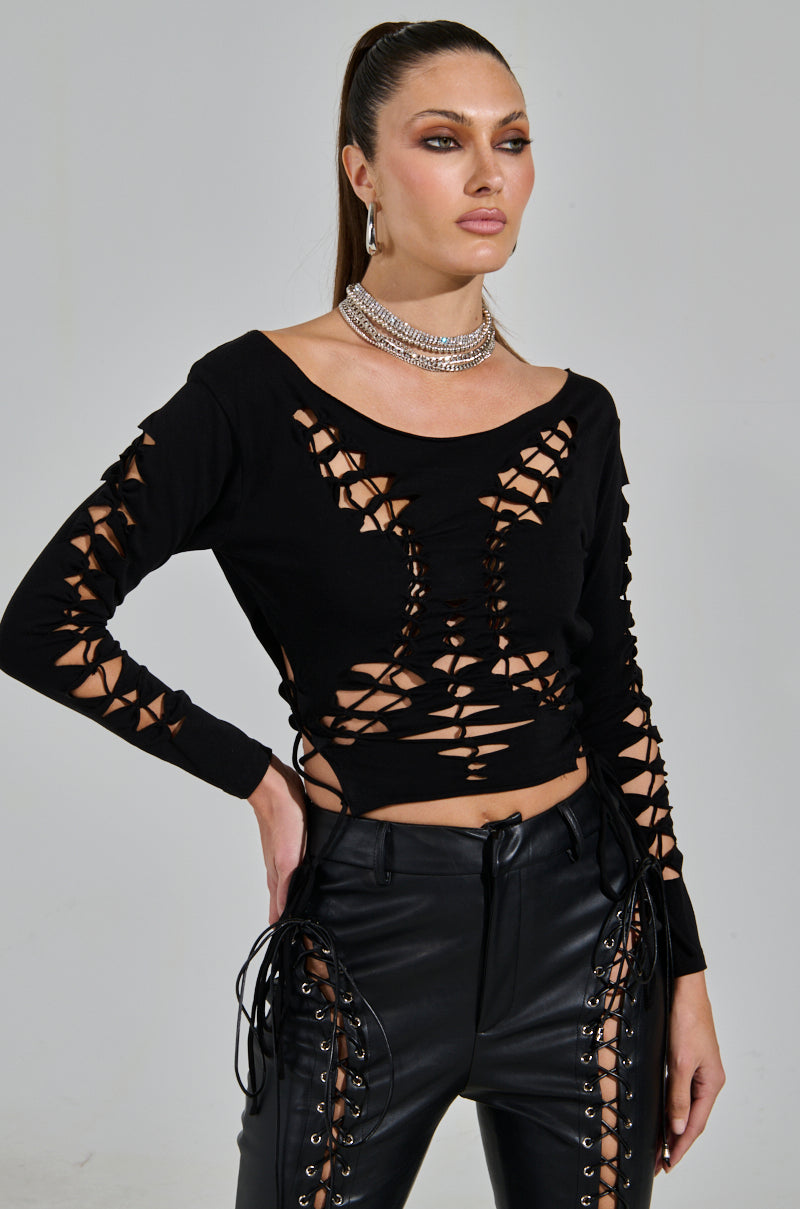A confident person poses in the RIP ME OUT DISTRESSED LONG SLEEVE TOP and leather pants featuring intricate lace-up details. With hoop earrings and a layered necklace, they exude glamour against a plain backdrop.