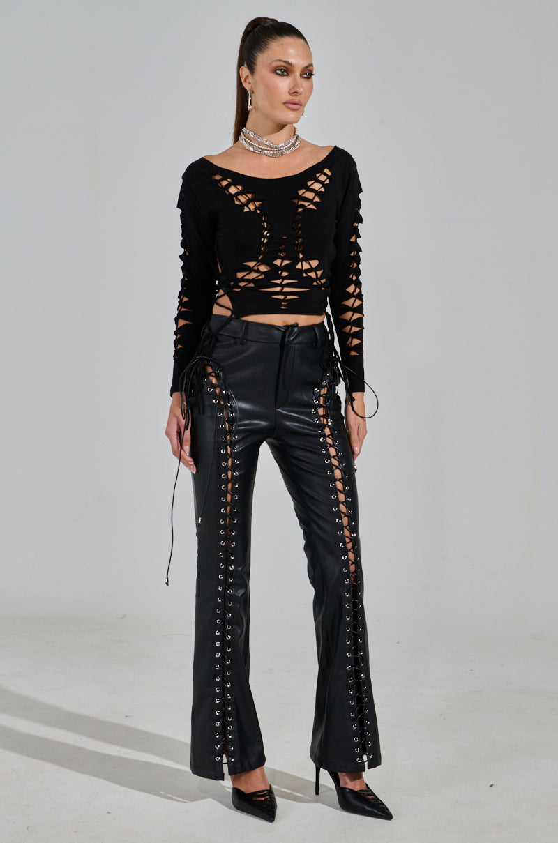 A model in the RIP ME OUT DISTRESSED LONG SLEEVE TOP and lace-up leather pants stands against a light backdrop. Her long hair is tied back, and she accessorizes with a necklace, earrings, and black pointed heels for an edgy elegance.