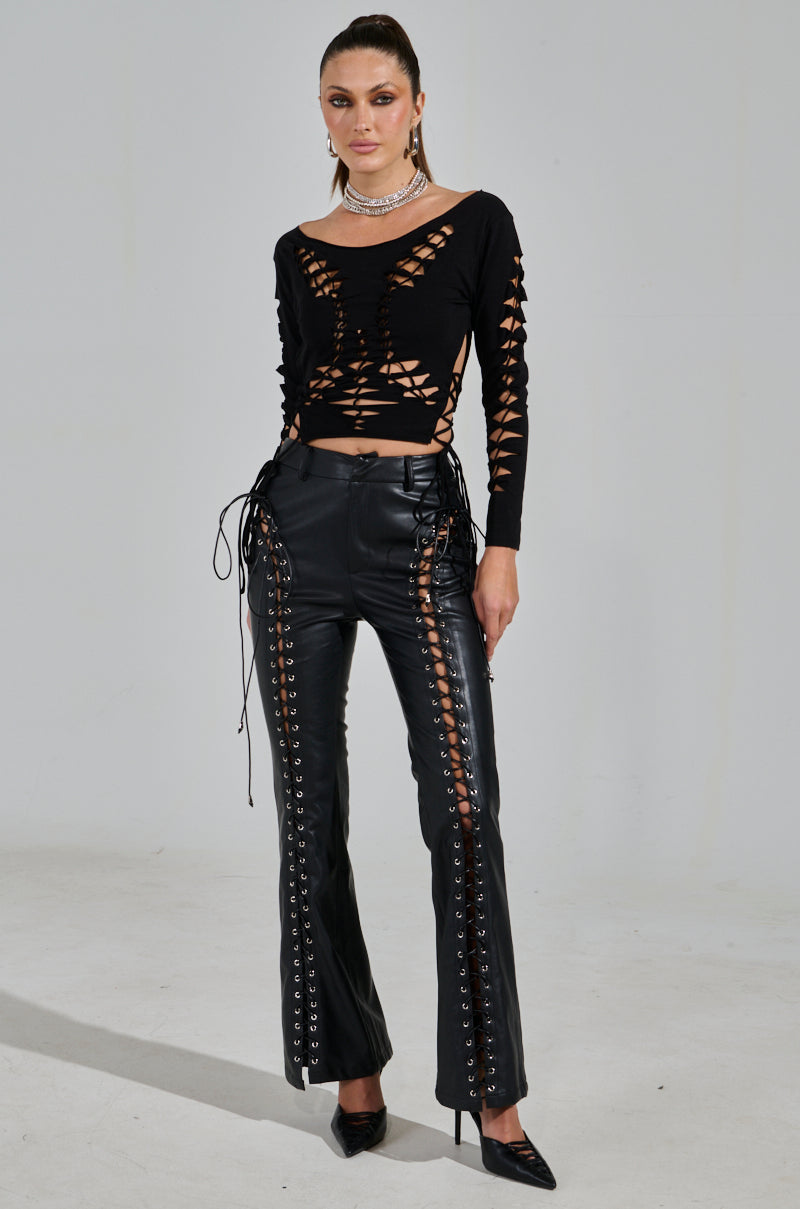 In a gray setting, someone exudes confidence in the RIP ME OUT DISTRESSED LONG SLEEVE TOP, paired with sleek black leather lace-up pants and chic black heels. Their hair is neatly tied back.