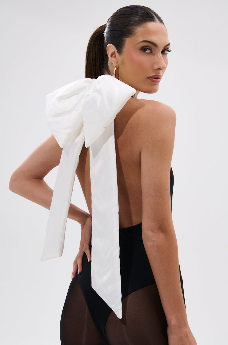 A woman with dark hair in a ponytail wears the LOVE ME MORE BOW BODYSUIT by AKIRA Label, a black body-contouring piece with a large white removable bow tied at the neck and long ribbon tails, posing against a plain light background. 0