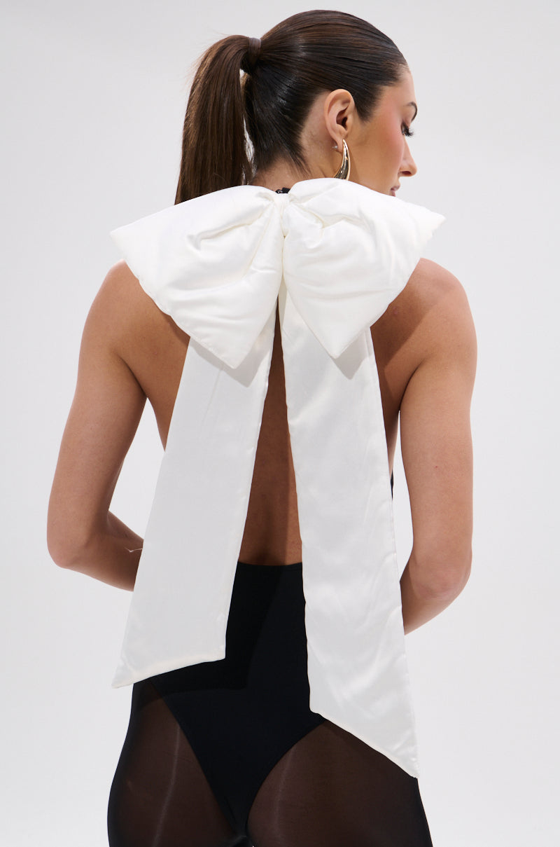 A woman with a sleek ponytail models the AKIRA Label LOVE ME MORE BOW BODYSUIT, a backless, body-hugging fit with a large removable white bow featuring long tails down her back. She faces away from the camera against a plain background. 3