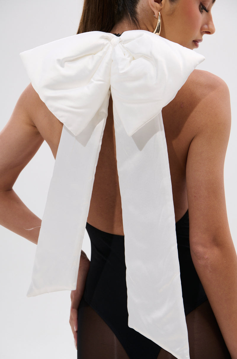 A woman in the LOVE ME MORE BOW BODYSUIT—a black, body-contouring piece by AKIRA Label—wears a large, removable white bow tied at her neck with long tails down her back. She accessorizes with gold hoops and has her hair pulled back. 7