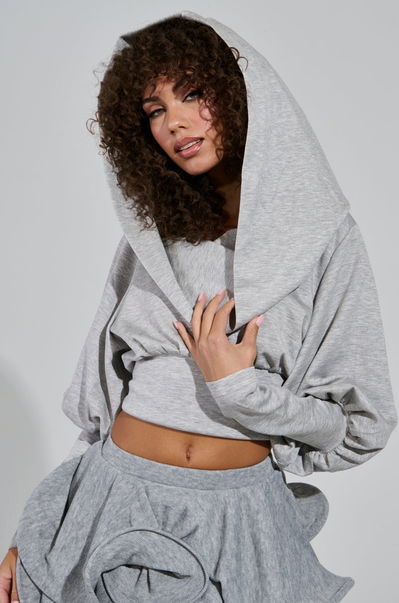 A woman with curly hair models the ATHENA HOODIE, highlighting a heather gray hooded ensemble paired with a ruffled skirt. The stretchy knit material drapes smoothly, while the cowl neckline falls gently over her head as she looks at the camera expressionlessly.