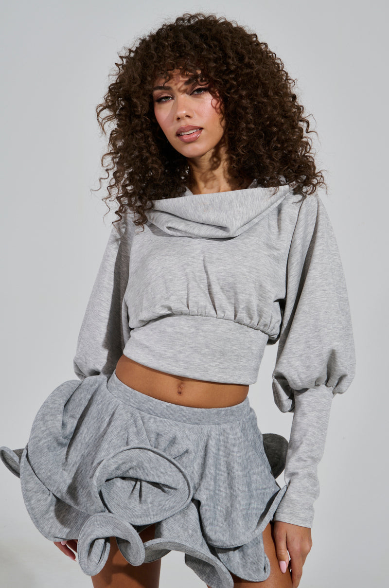 Wearing a matching gray ensemble, a person with curly hair dons the ATHENA HOODIE featuring a cowl neckline and pairs it with a skirt adorned with large, sculptural ruffles. Crafted from stretchy knit fabric, they stand against a plain white background.