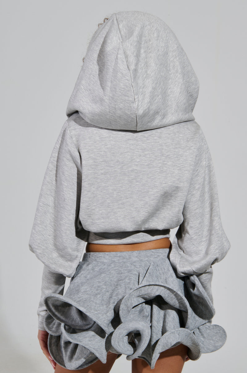 A person clad in the ATHENA HOODIE featuring a cowl neckline, paired with a distinctive gray skirt adorned with ruffled layers, stands facing away from the camera. The contemporary and artistic ensemble creates a striking contrast against the simple background.