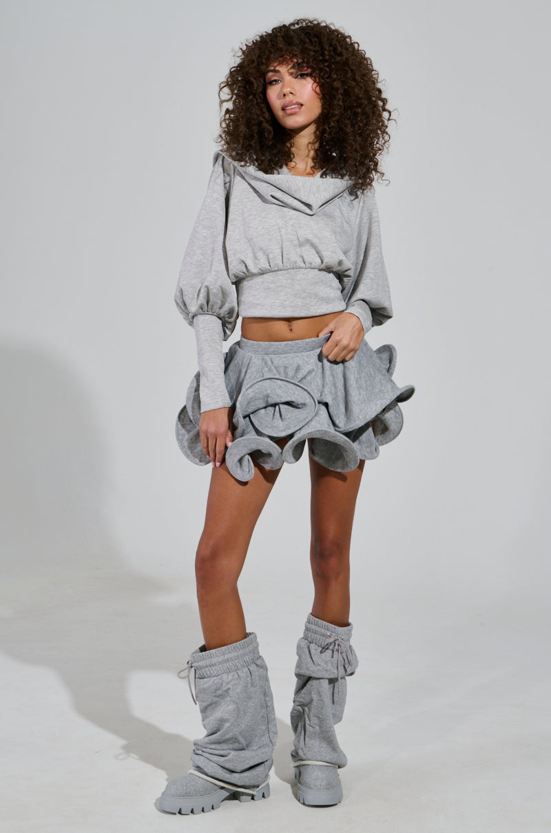 A woman with curly hair showcases a distinctive gray ensemble, featuring the ATHENA HOODIE with its cowl neckline and a ruffled mini skirt. Her matching gray slouchy leg warmers and shoes complete the look as she poses against a plain backdrop.