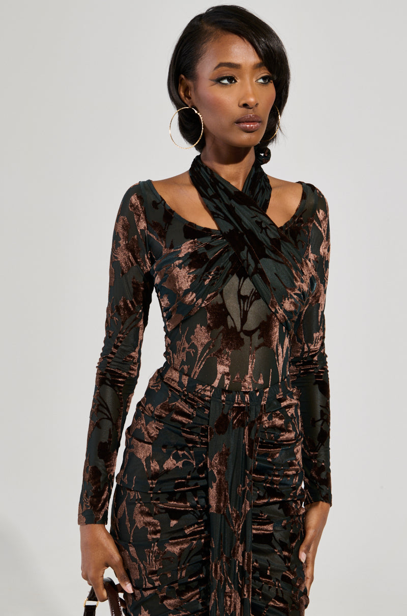 A woman in the Always In The Mood bodysuit, featuring a fitted long-sleeve floral velvet pattern in dark tones, stands against a plain backdrop. She accessorizes with hoop earrings and sports a side-swept hairstyle.