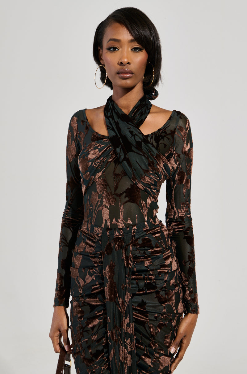 A woman stands confidently in an Always In The Mood Bodysuit in brown, featuring a unique velvet floral pattern. Her hair is neatly styled, she wears hoop earrings, and the background is a simple light color.