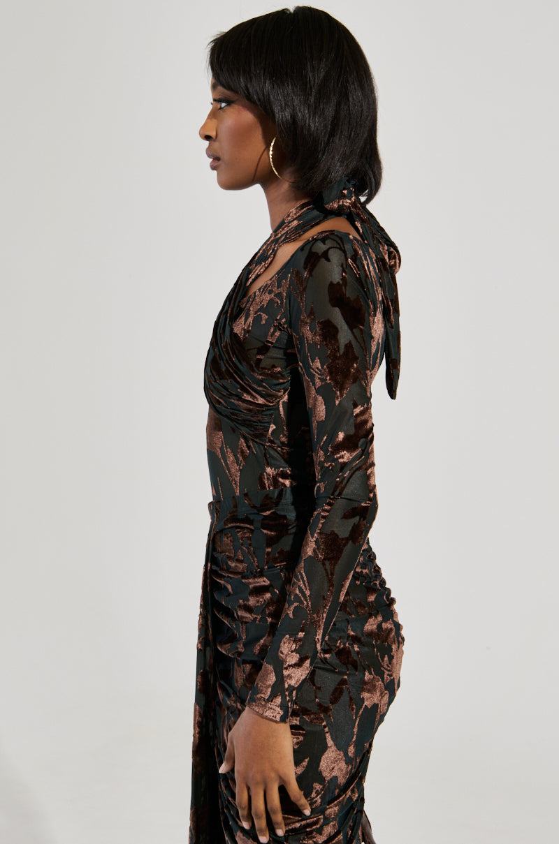 A woman in a form-fitting, long-sleeve Always in the Mood Bodysuit in Brown with a shimmering velvet floral pattern stands sideways. Her shoulder-length dark hair and large hoop earrings contrast elegantly against the plain, light-colored background, highlighting her silhouette.