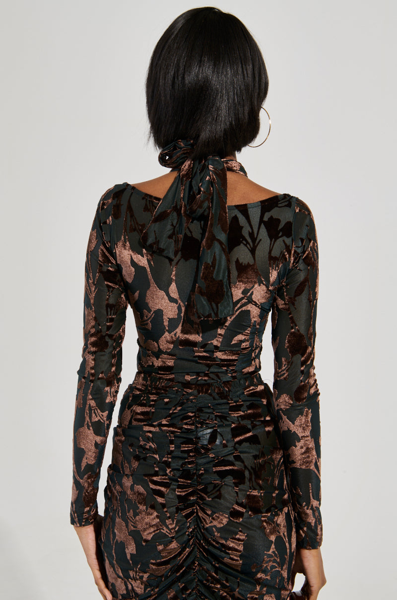 Rear view of a person with shoulder-length dark hair wearing the Always In the Mood Bodysuit in Brown, featuring a fitted long-sleeve design with a sheer, velvet floral pattern, accented by a tied bow at the neck and ruching down the back.
