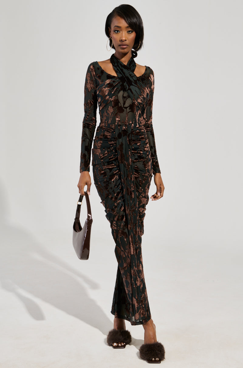A person in a long, patterned dress flaunts the ALWAYS IN THE MOOD BODYSUIT in brown with velvet floral accents and a tie detail. They carry a dark purse, their short hair complements furry shoes beautifully against a light, plain background.
