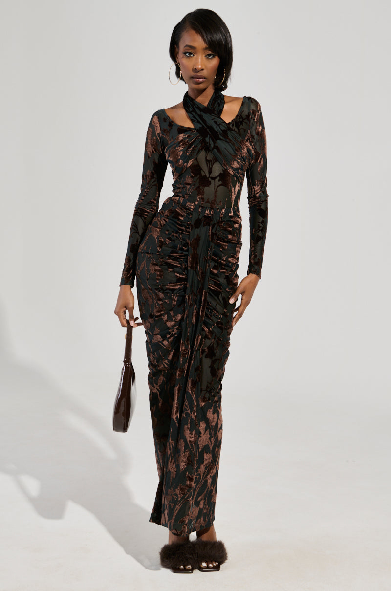 Dressed in the "ALWAYS IN THE MOOD BODYSUIT IN BROWN," a figure showcases an elegant, form-fitting outfit with a sheer mesh overlay and high neckline featuring a velvet floral pattern. They hold a small, dark handbag and wear furry sandals against a plain, light background.