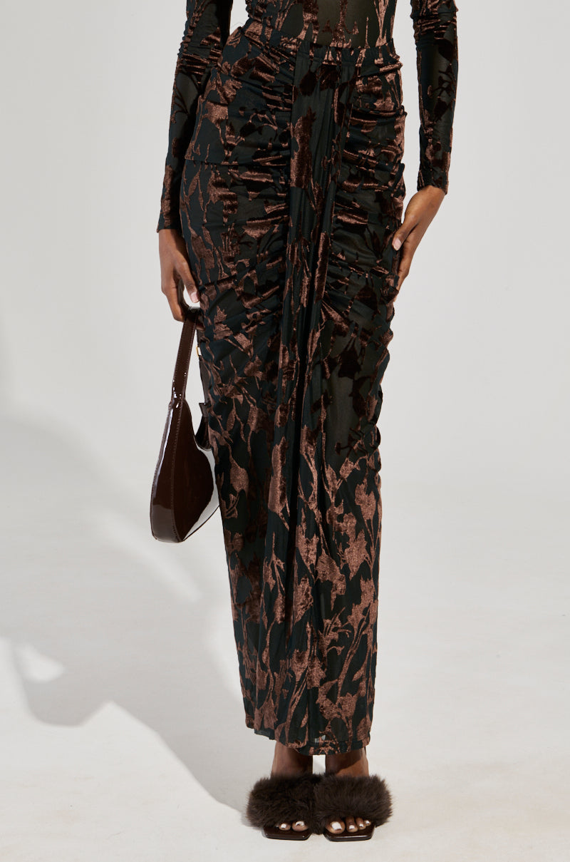 A person wearing the ALWAYS IN THE MOOD MAXI SKIRT, featuring a curve-hugging silhouette with a black and brown floral pattern, holds a dark handbag and pairs it with fluffy, open-toed shoes. The backdrop is set in a simple, light-colored studio setting.