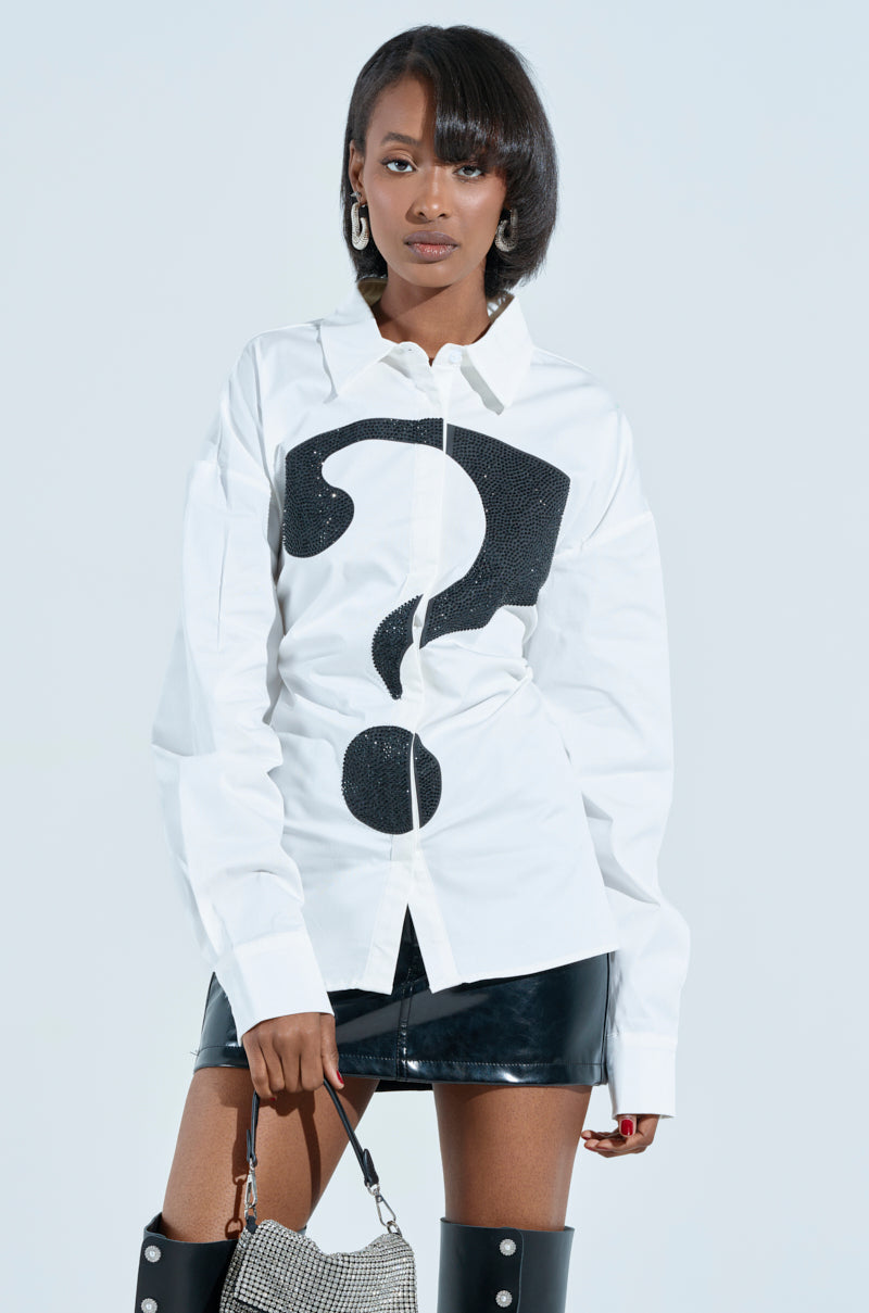 A woman confidently wears a RHINESTONED QUESTION BUTTON UP featuring a bold black question mark on white. She pairs it with a black leather skirt, holds a small handbag, and completes the look with hoop earrings and straight shoulder-length hair against a plain backdrop.