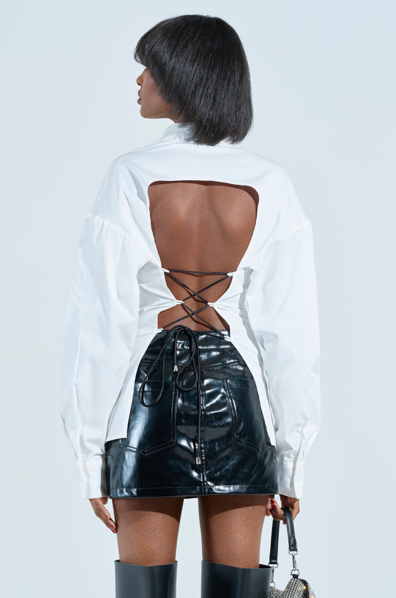 Clad in the RHINESTONED QUESTION BUTTON UP with its open-back, crisscross lace design, and shiny black leather skirt, a person stands facing away. They hold a small handbag and wear tall black boots, channeling an AKIRA Label vibe against a light gray backdrop.