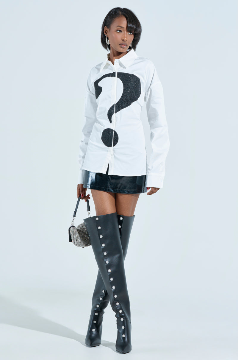 A person confidently wears the RHINESTONED QUESTION BUTTON UP with a black leather skirt, knee-high black buttoned leather boots, and carries a small handbag. Their short, styled hair complements the exclusive AKIRA Label piece against a light gray background.