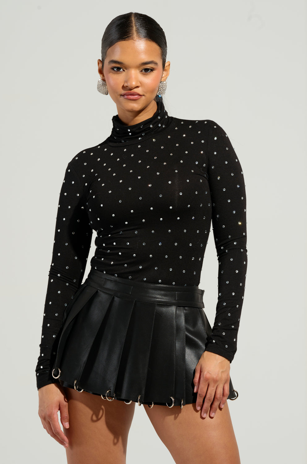 A person wearing the SPARKLING TURTLENECK TOP, a black long-sleeve knit top with rhinestone embellishments, and a black pleated leather skirt stands against a plain background. They have long earrings and their hair is tied back.