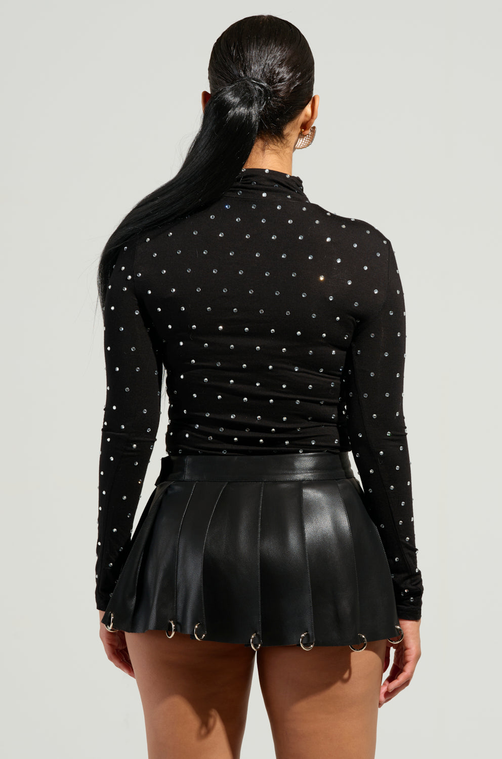 A person with long dark hair is styled in the SPARKLING TURTLENECK TOP paired with a black pleated leather skirt featuring ring details. Rhinestone embellishments add an extra touch of elegance to their captivating ensemble as they face away from the camera.
