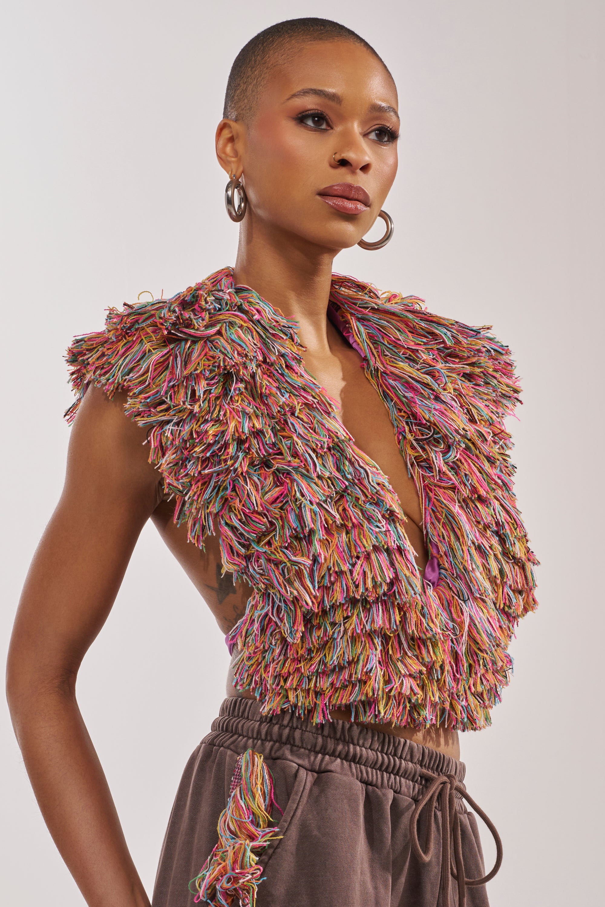 A person with a shaved head wears hoop earrings, high-waisted brown pants, and THE ONE FRINGE BLOUSE—a sleeveless, multicolored shaggy-textured top that makes a bold statement against a plain background. 0