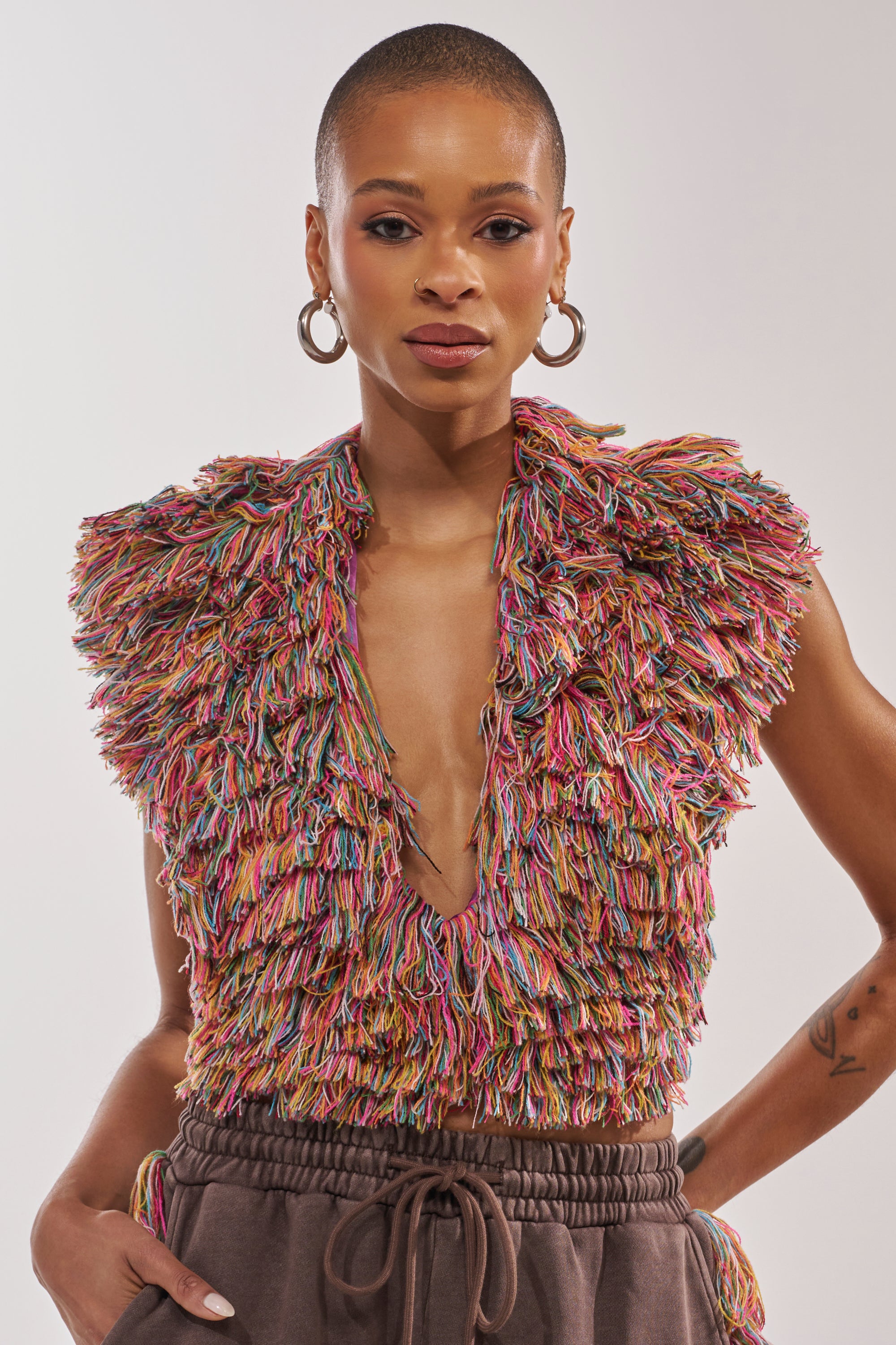 Wearing THE ONE FRINGE BLOUSE with a deep V-neck, large hoop earrings, and brown drawstring pants, a person with short hair confidently poses against a plain background, their statement layering adding unique flair. 1