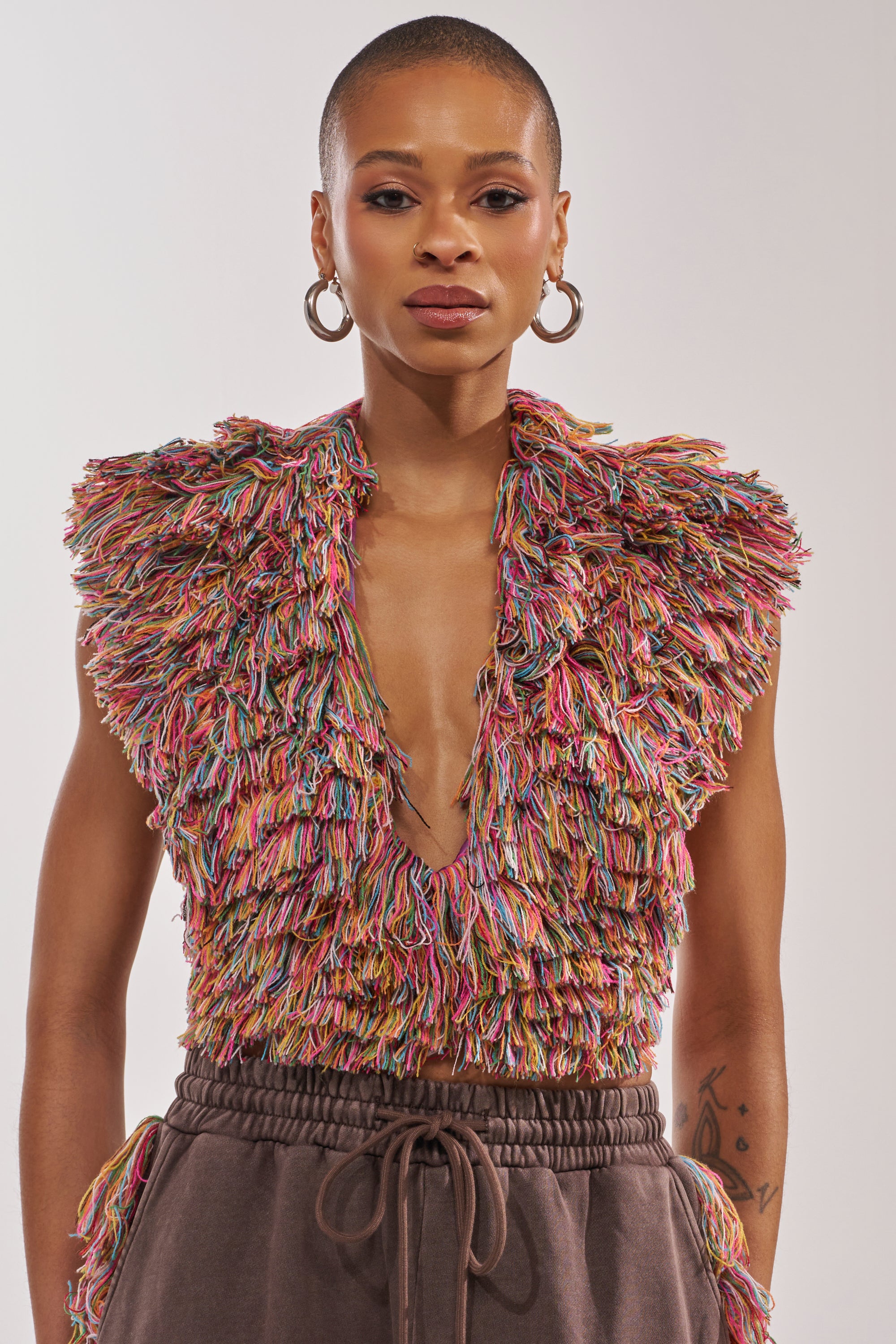 A person with short hair and hoop earrings models THE ONE FRINGE BLOUSE, a colorful rainbow cropped top with a deep V-neck, paired with brown drawstring pants—ideal for festival wear—against a plain background. 2