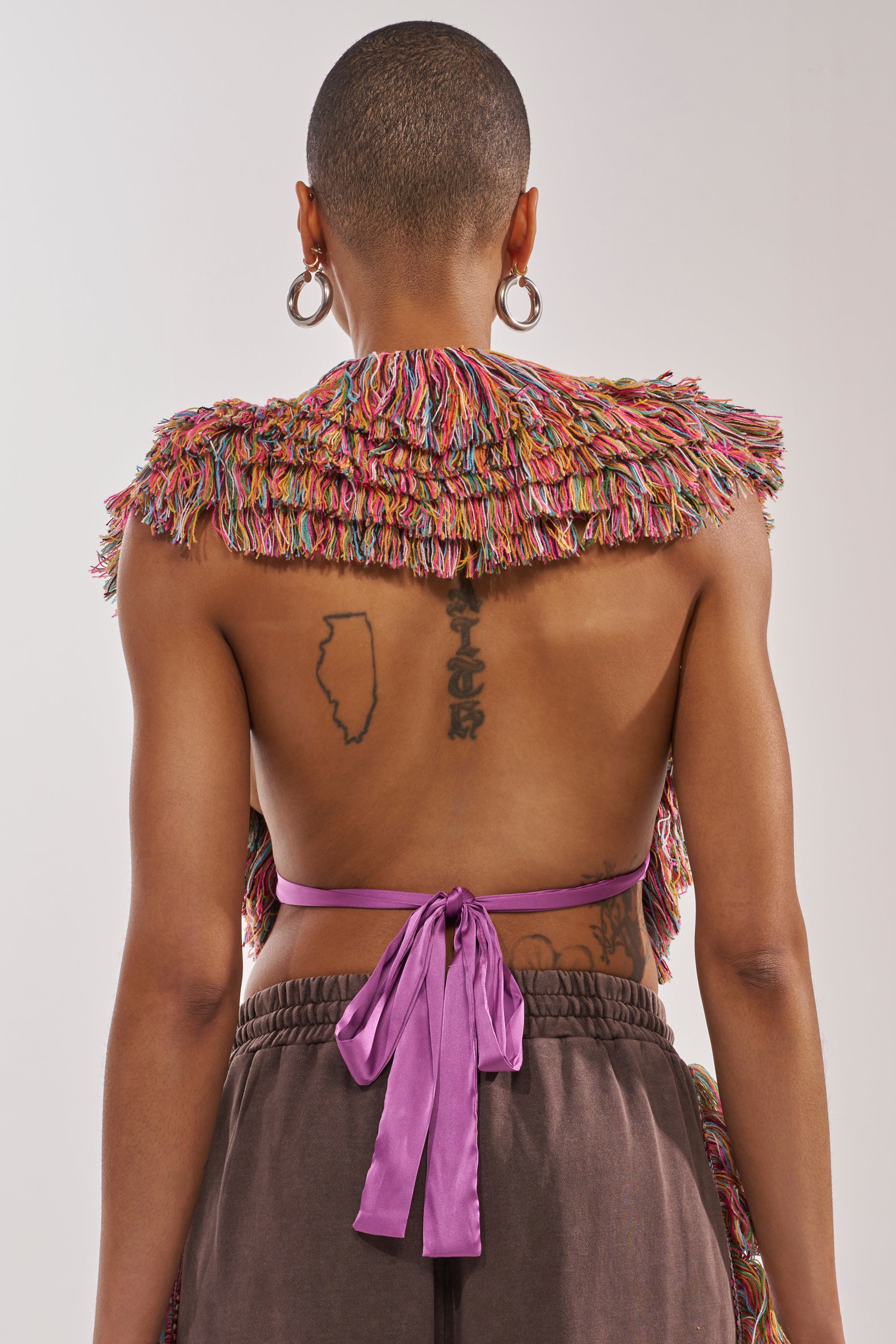 Wearing THE ONE FRINGE BLOUSE—a colorful, textured, backless top with a purple ribbon—paired with brown pants and bold accessories, this look is perfect for festivals and makes an eye-catching fashion statement. 3