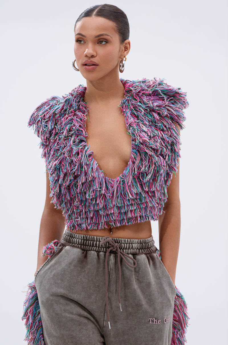 A woman wears THE ONE FRINGE BLOUSE, a multicolor fringe top with a deep V-neck, paired with loose brown drawstring pants. With hands in her pockets, she exudes editorial loungewear style against a plain light background.