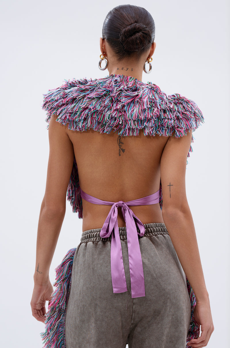 A person with a bun showcases editorial style in THE ONE FRINGE BLOUSE—a multicolor fringe crop top with a large lavender ribbon—paired with high-waisted gray pants. Tattoos decorate their arms and upper back.