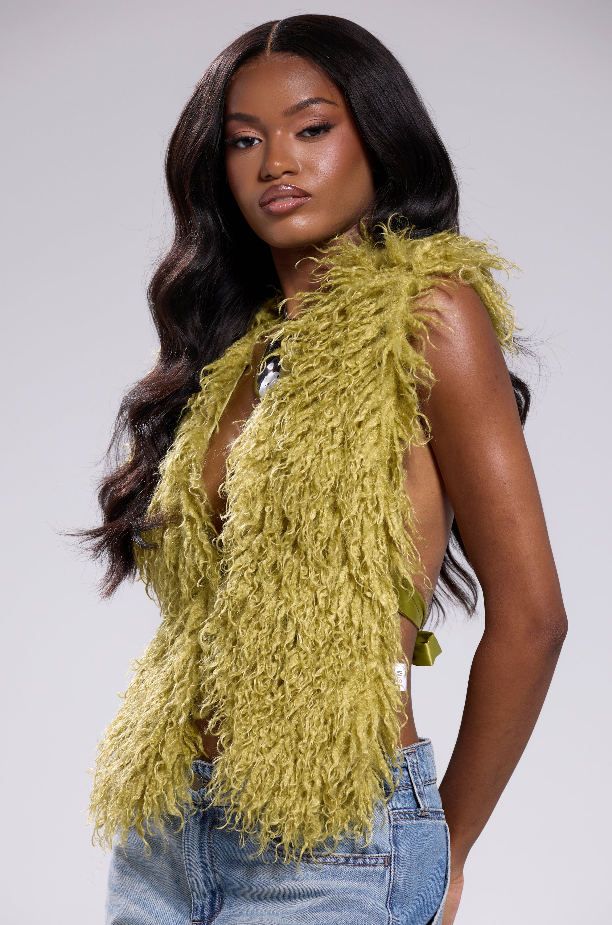 A woman with long, wavy dark hair models the JAMEELA TOP IN GREEN, a sleeveless lime green faux fur style, paired with blue jeans. She poses confidently against a plain light background, looking directly at the camera.