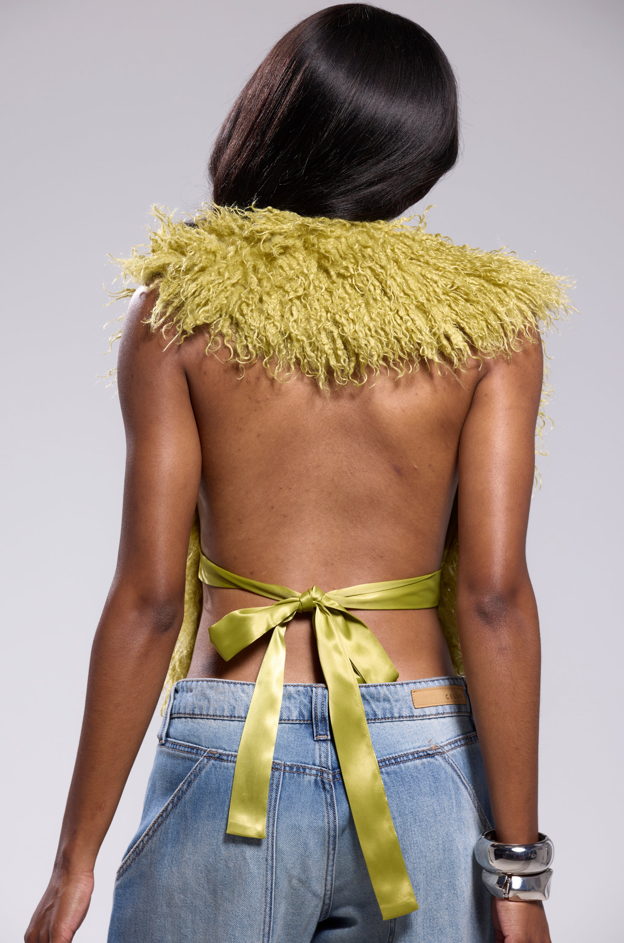 A person with straight dark hair wears the JAMEELA TOP IN GREEN, a lime green textured backless top tied with a satin ribbon, paired with loose blue jeans and a chunky metallic bracelet, standing with their back to the camera.