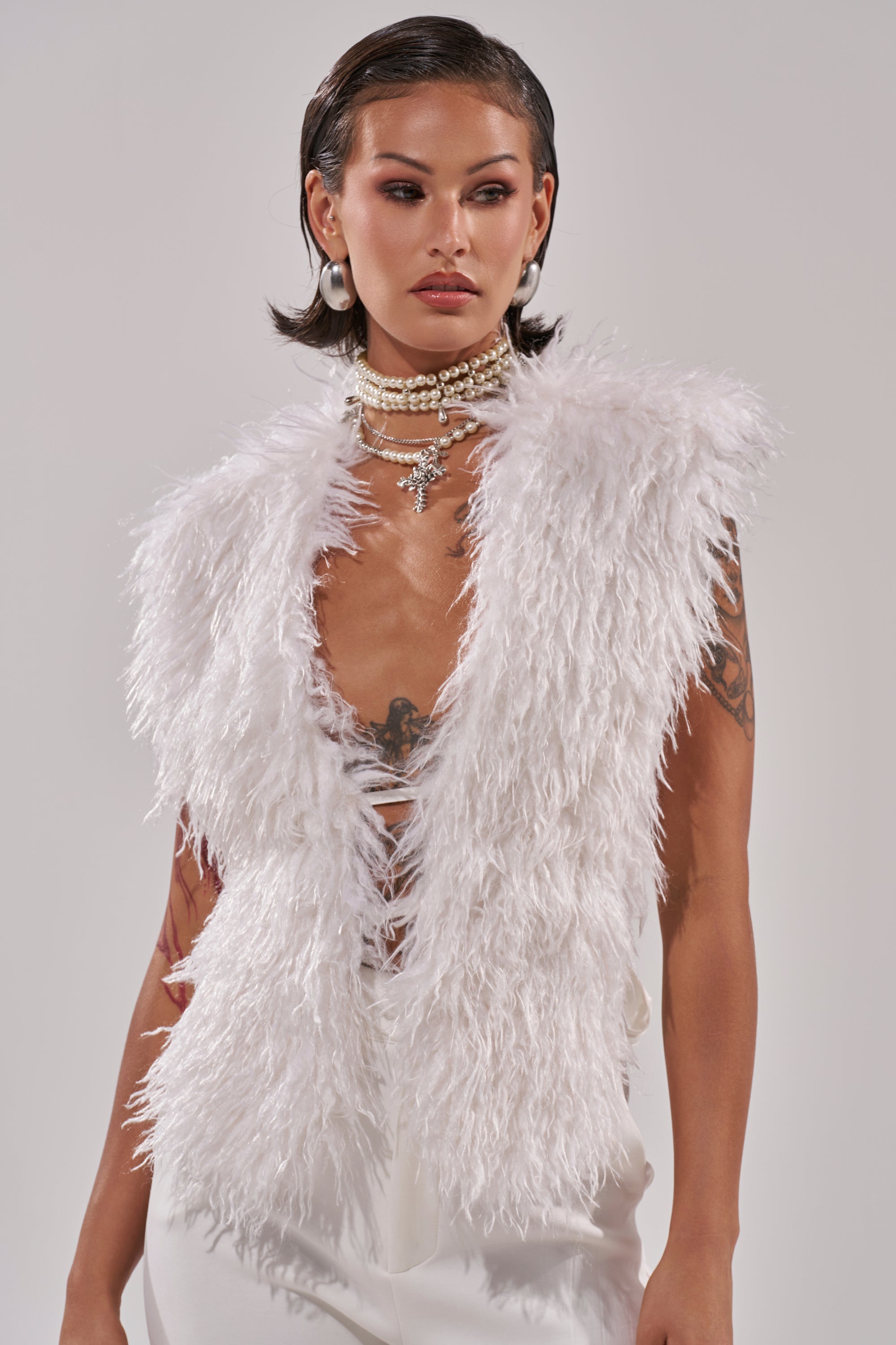 A person with short dark hair and visible arm tattoos poses against a plain light background wearing the JAMEELA FAUX FUR TOP, white pants, large hoop earrings, and layered pearl necklaces.