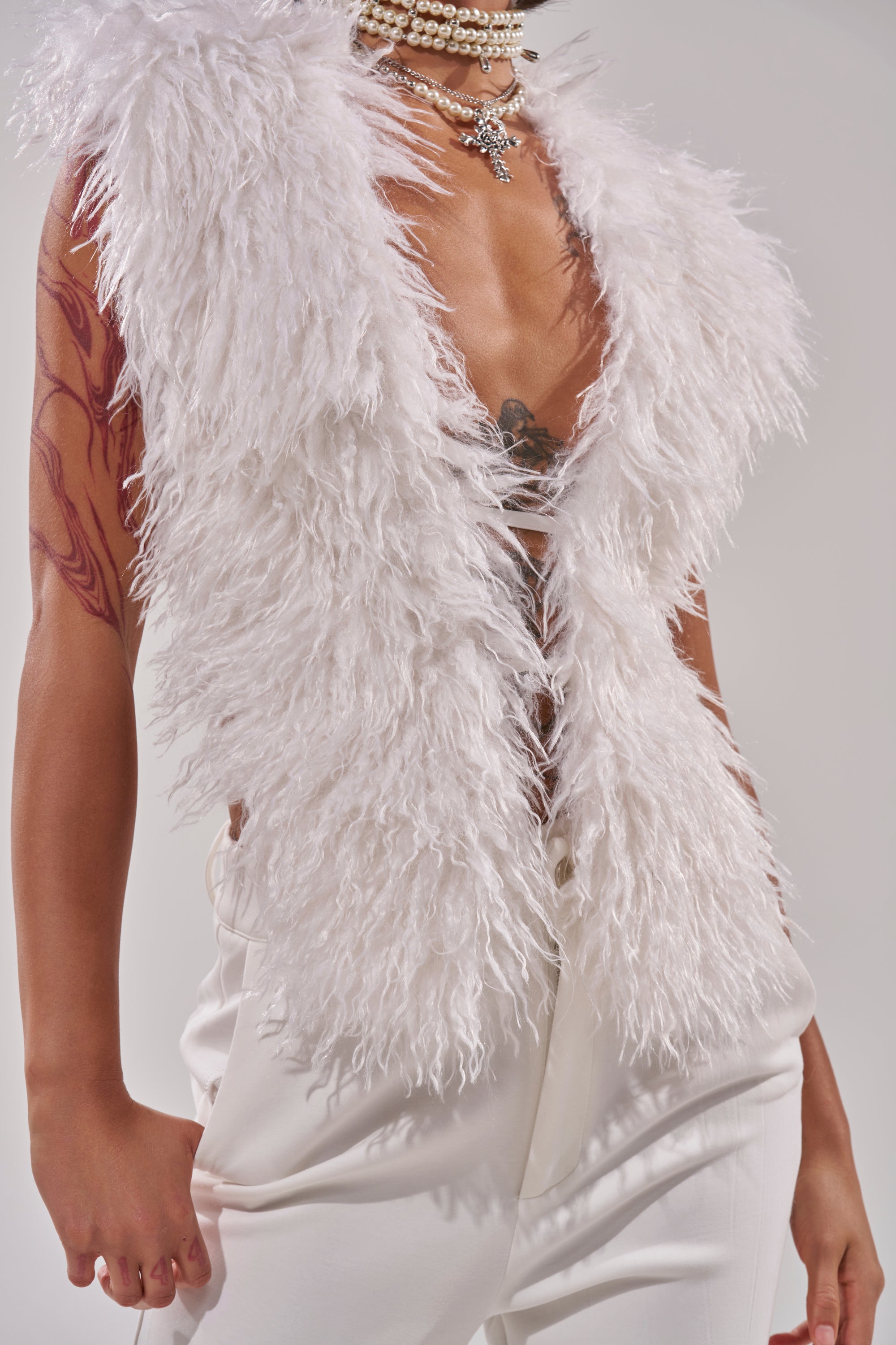A person in the JAMEELA FAUX FUR TOP, white pants, layered pearl necklaces with a star pendant and satin ribbon, shows off a tattooed arm against a plain light background.