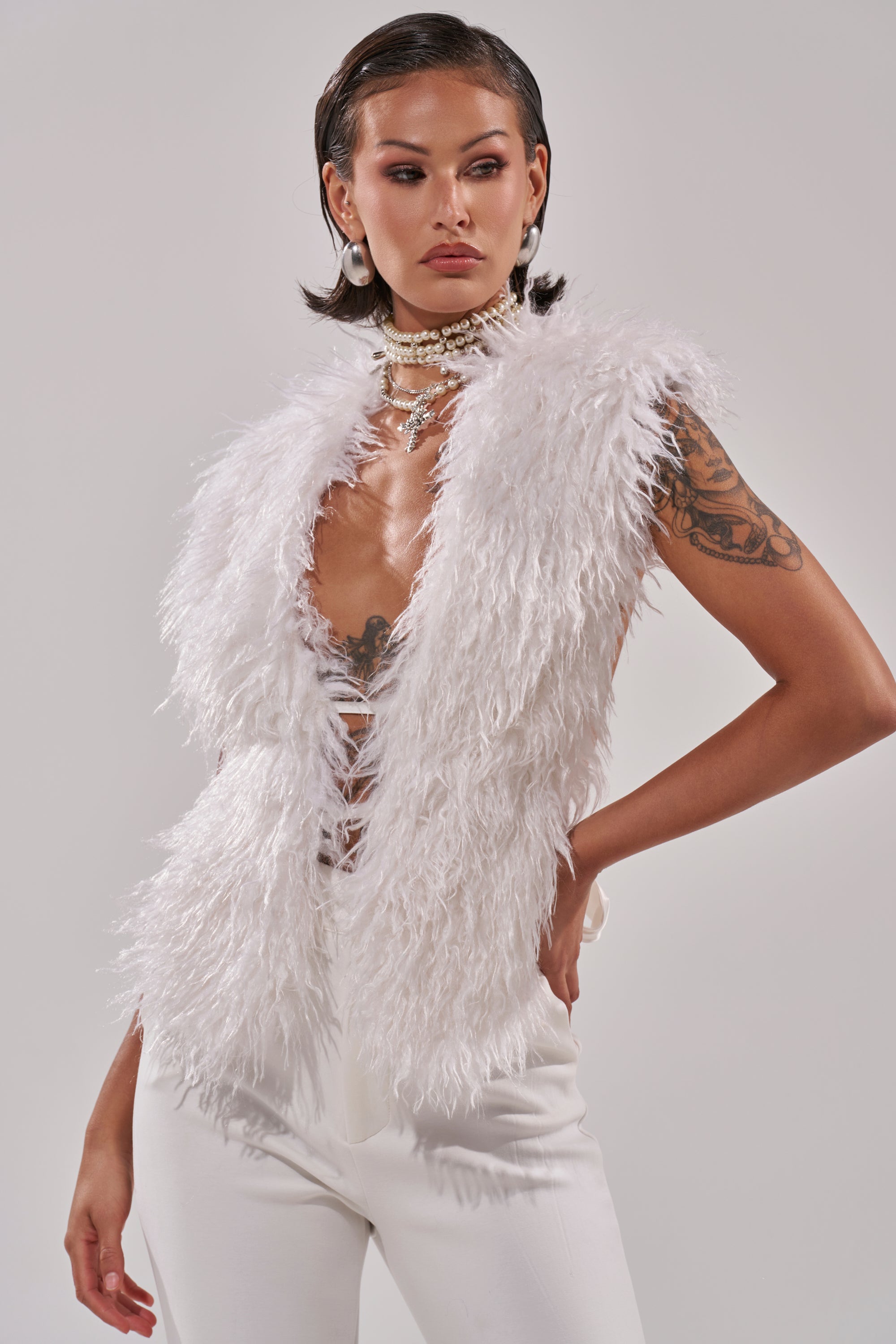 A woman with short, slicked-back hair and visible tattoos poses confidently against a plain background wearing the JAMEELA FAUX FUR TOP in ivory, paired with white pants, pearl earrings, and layered pearl necklaces.