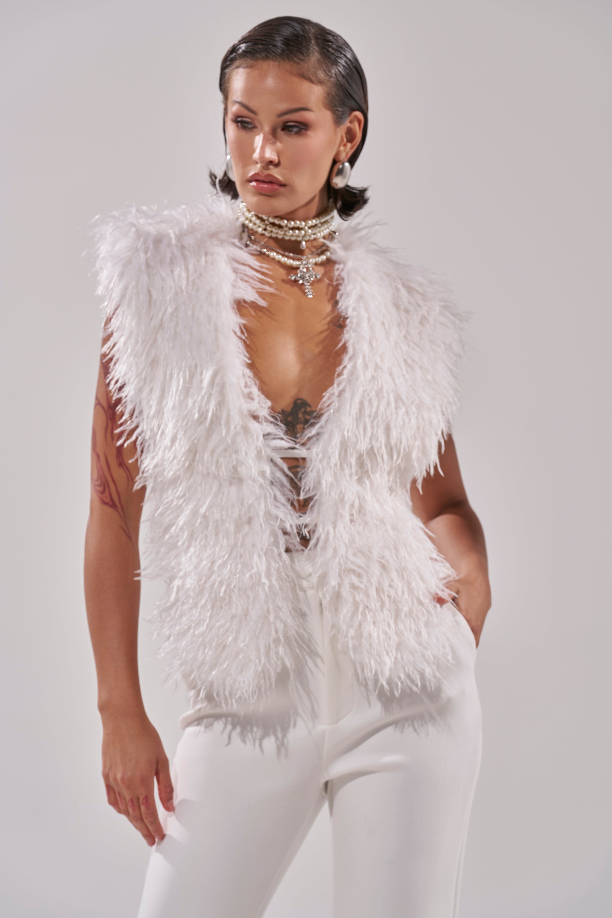 A person wears the JAMEELA FAUX FUR TOP in ivory with white pants, layered pearl necklaces, and a sheer top, posing with one hand in their pocket against a plain background.