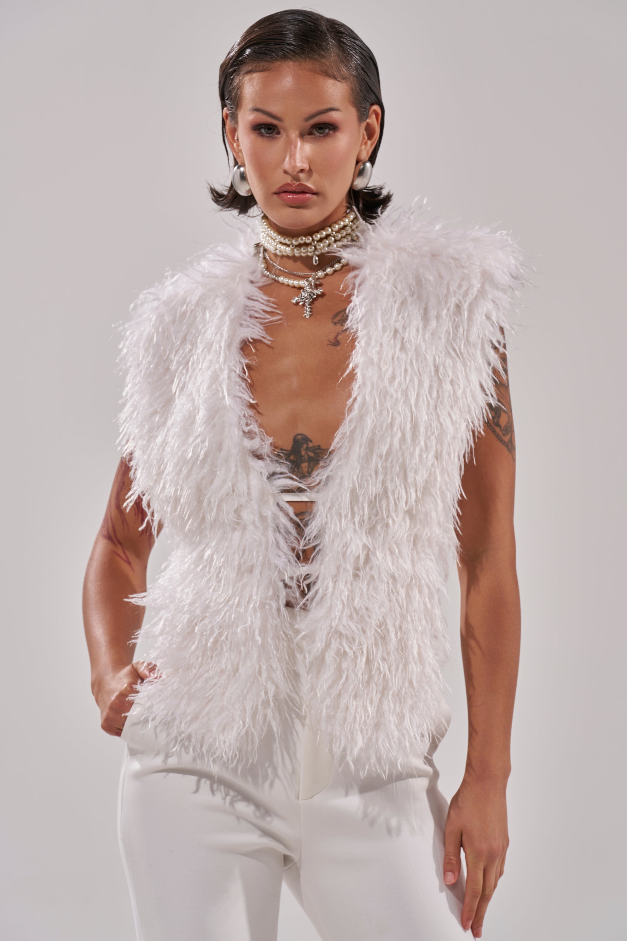 A person with slicked-back short dark hair models the ivory JAMEELA FAUX FUR TOP over white pants, accessorized with layered pearl necklaces and visible tattoos, standing against a plain light background.